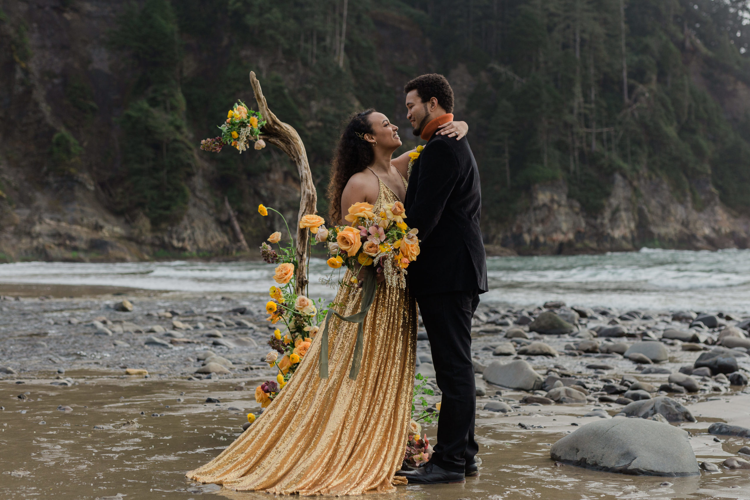 A bride wearing a gold wedding dress is wrapping her hands around her partner in the coast of Oregon. The bride is holding a bouquet on her other hand.